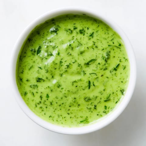 Bright green Green Goddess Dressing in a glass jar, garnished with chopped chives and parsley, sitting beside crisp lettuce leaves.